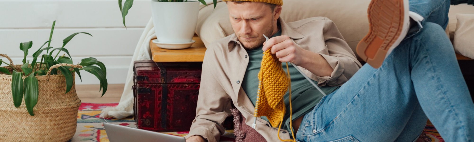 Young serious man looking at laptop. Man learning new hobby, knitting on needles. Knitting project in progress. - Image